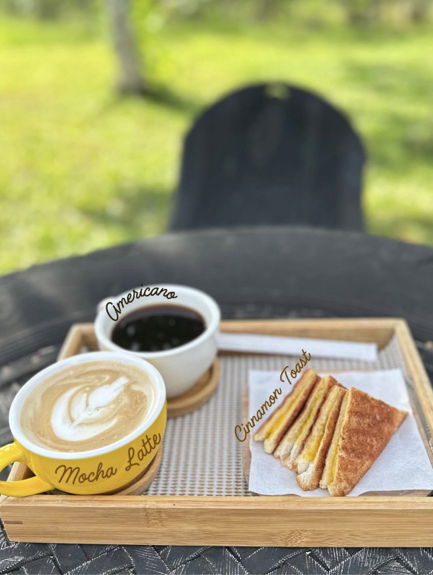 A wooden tray on a garden chair holding an Americano, a yellow mug of Mocha Latte with leaf latte art, and a plate of sliced cinnamon toast