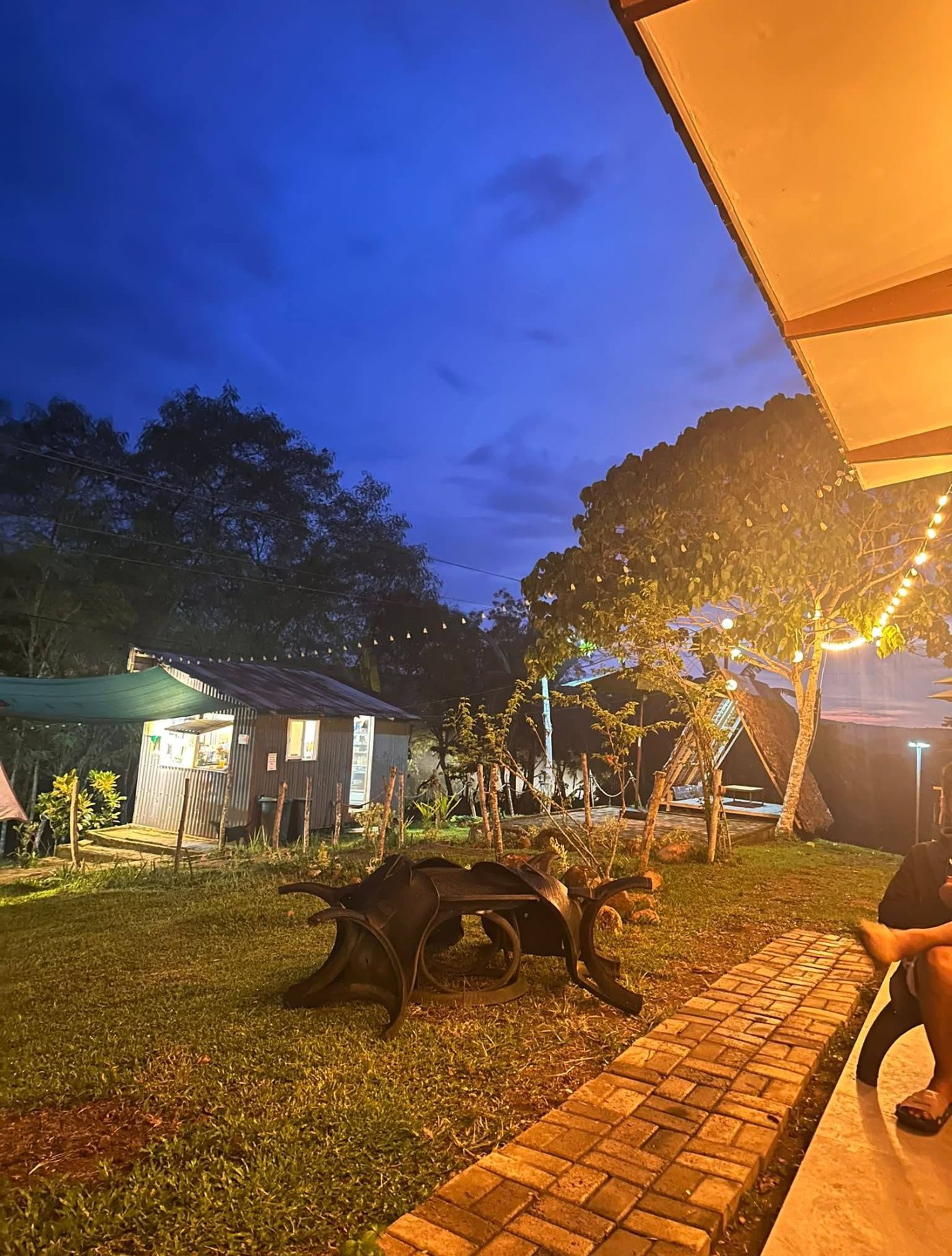 The Lawagon grounds at blue hour — the service hut on the left, an A-frame shelter under the trees, and a brick path running between them