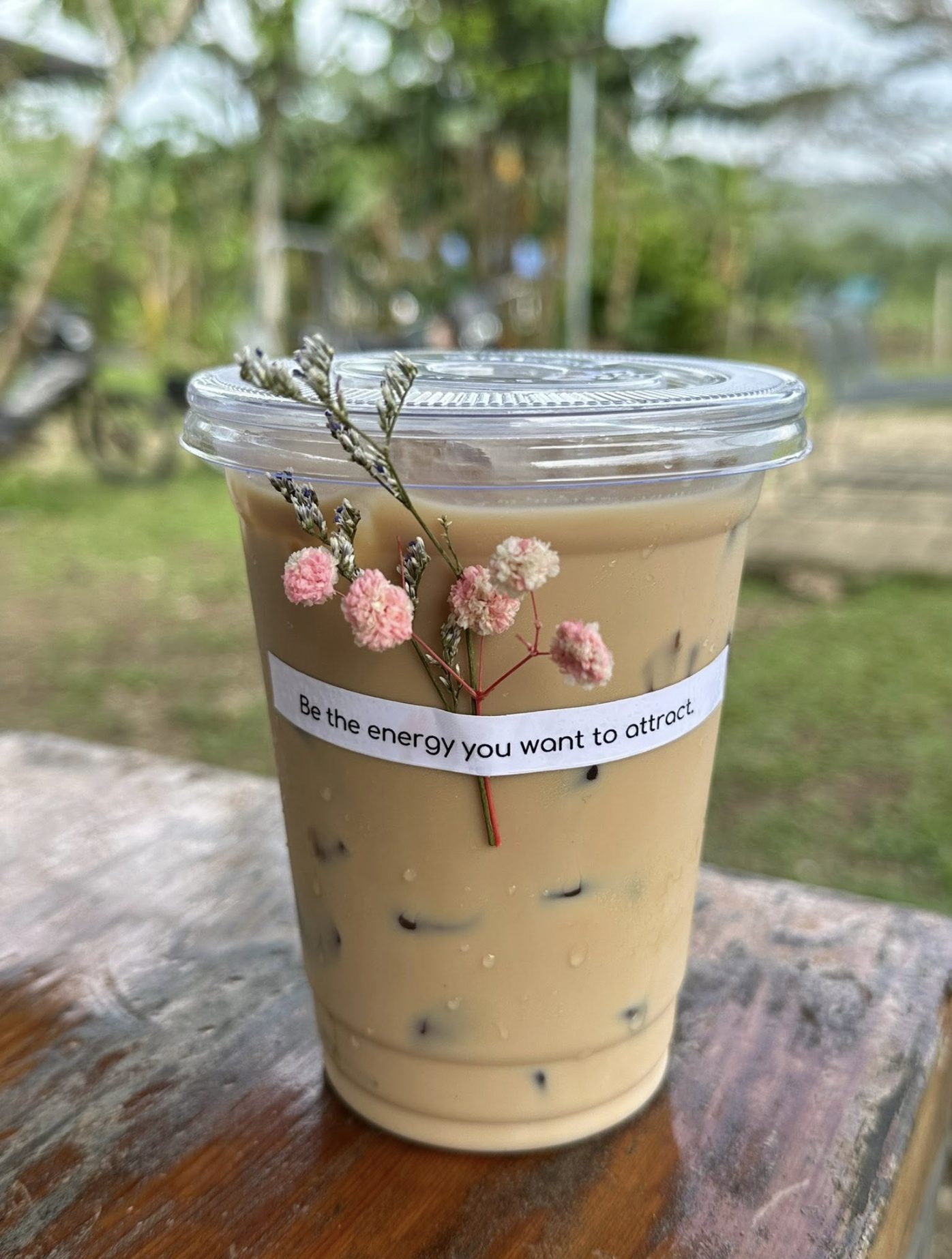 An iced latte in a clear cup on a wooden rail, dried flowers tucked against a paper strip reading “Be the energy you want to attract.”