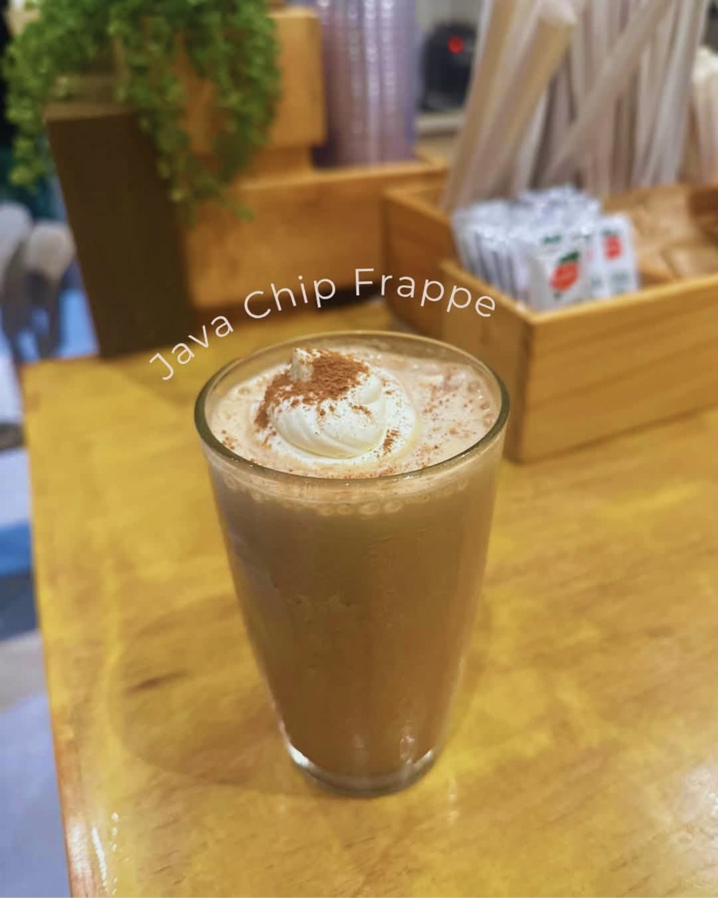 A Java Chip Frappé in a tall glass, topped with whipped cream and a dusting of cinnamon, on a pale wooden table