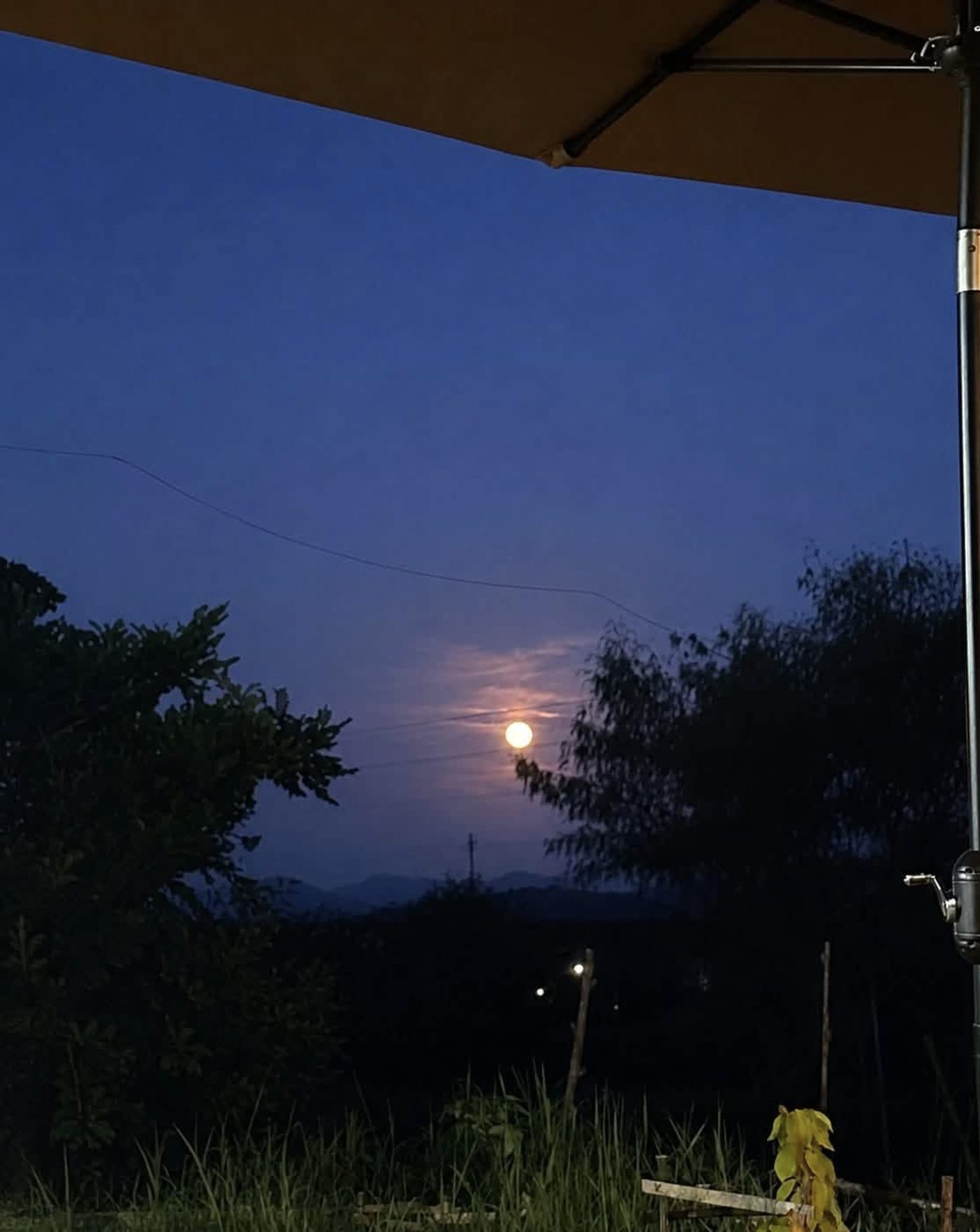 A full moon rising over distant hills, seen from under a canvas umbrella at the edge of the garden