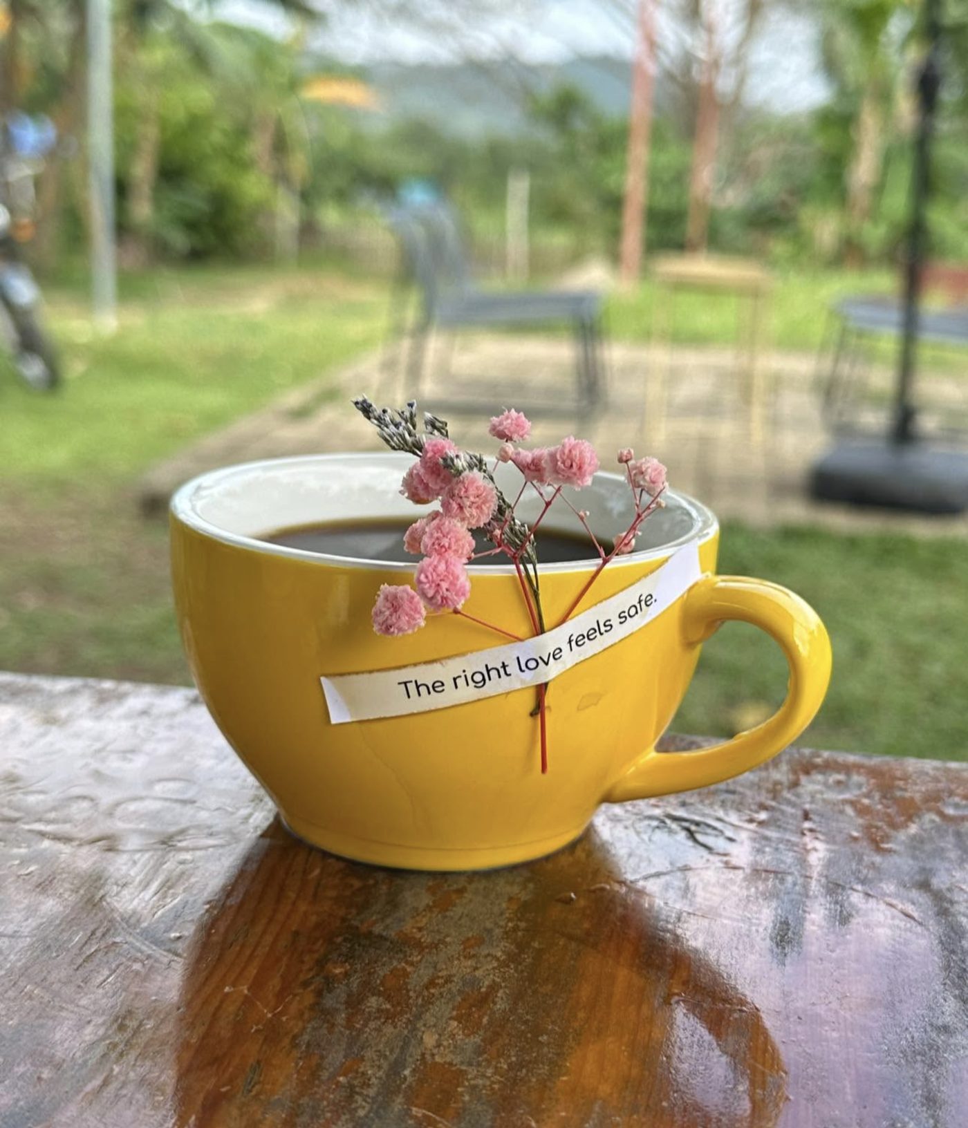 A yellow ceramic mug of black coffee on a wooden ledge, a sprig of dried pink flowers laid across it and a paper strip reading “The right love feels safe.”